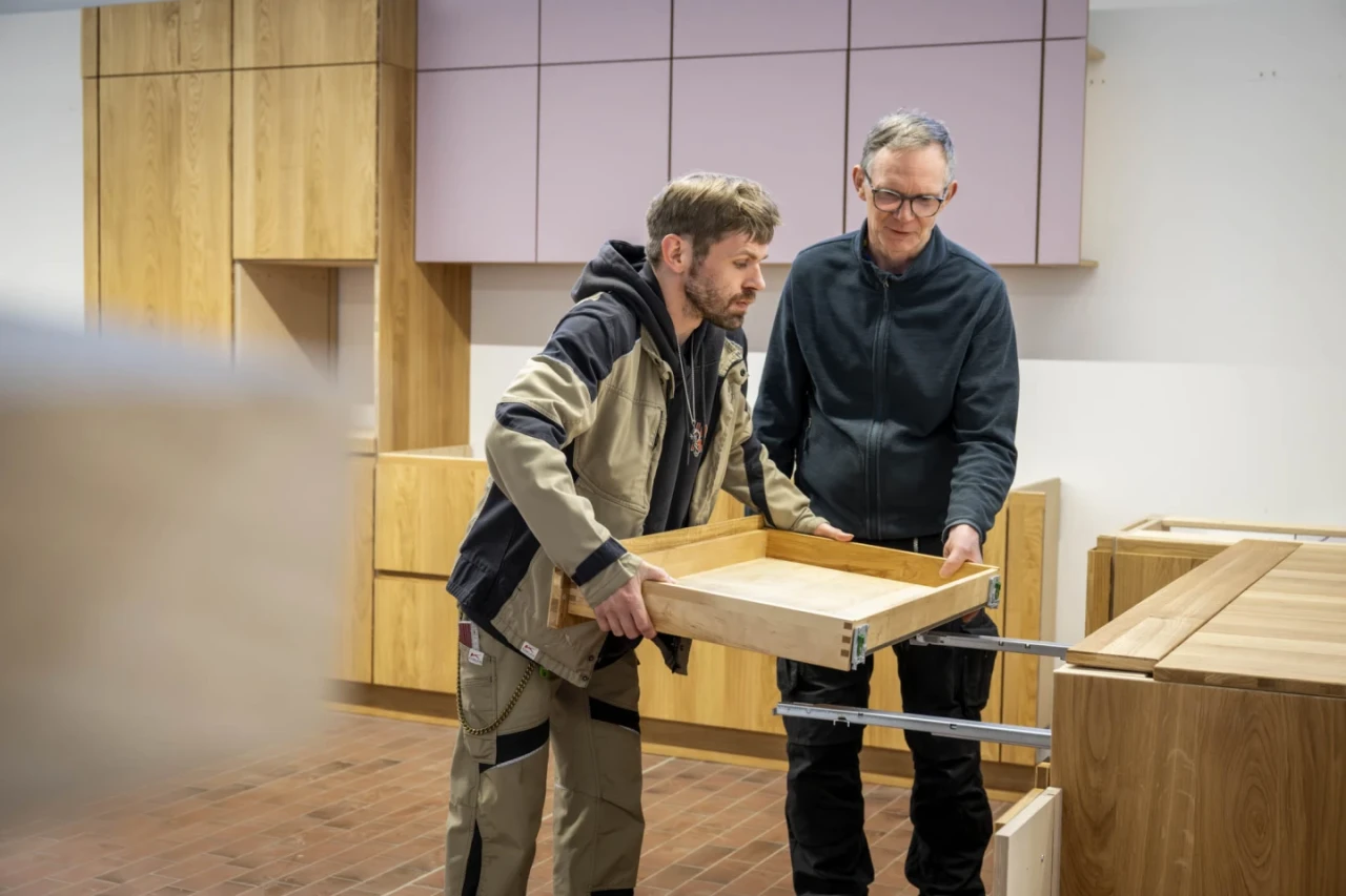 Zwei M&auml;nner in einer Holzwerkstatt montieren eine Schublade. Links ein Mann in Arbeitskleidung mit brauner Jacke und beiger Hose, rechts ein Mann mit Brille und dunklem Pullover. Im Hintergrund Holzschr&auml;nke mit lila Elementen an einer wei&szlig;en Wand. Boden aus roten Ziegeln.