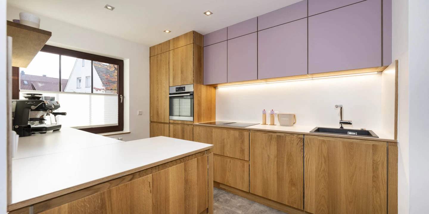 Moderne K&uuml;che mit h&ouml;lzernen Unterschr&auml;nken und lila-gr&auml;ulichen Oberschr&auml;nken. Links K&uuml;chenzeile mit Kaffeemaschine und Fenster mit Blick auf D&auml;cher. Mittig eingebauter Backofen, rechts Sp&uuml;le, daneben Arbeitsplatte mit zwei Gew&uuml;rzdosen. Grauer Boden und wei&szlig;e Decke mit Einbauleuchten.