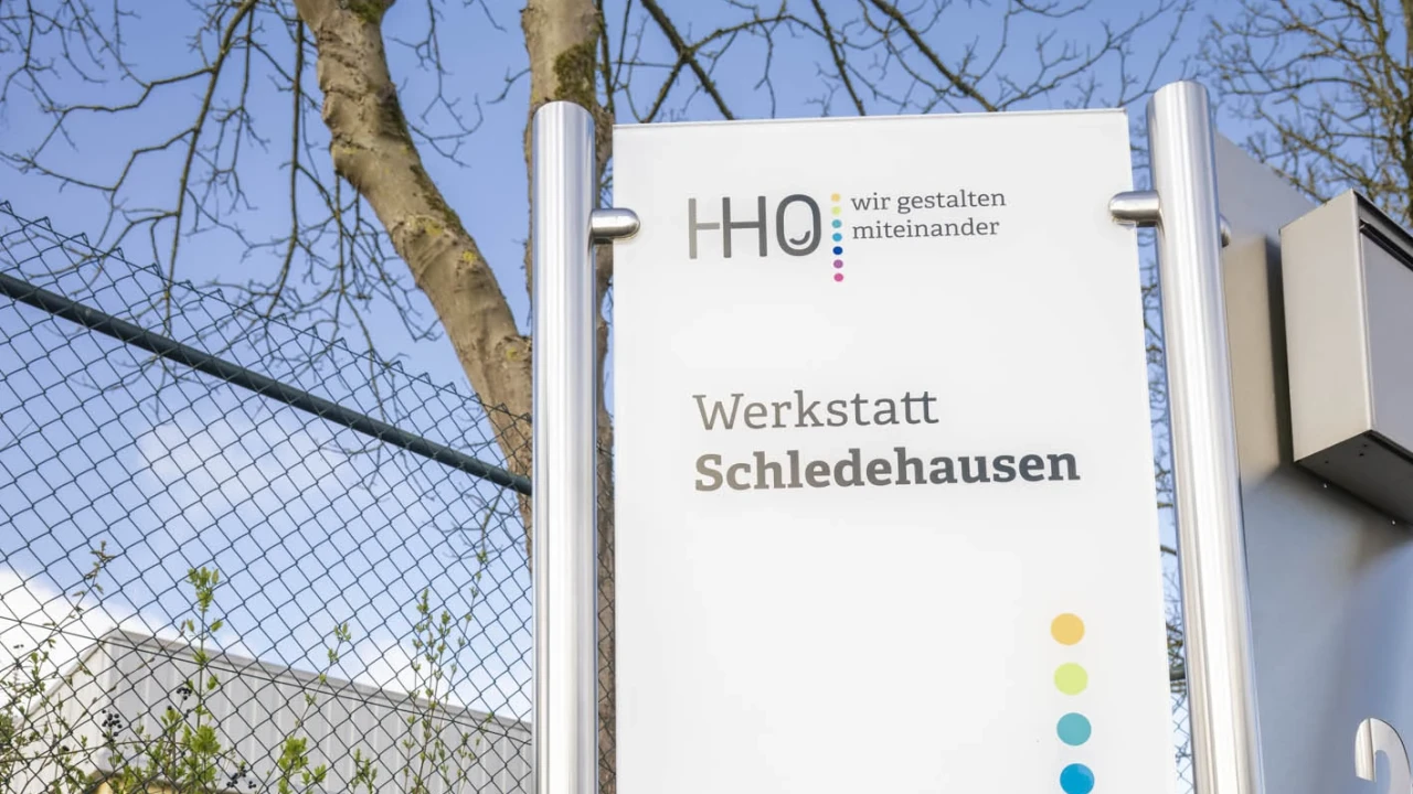 Rechteckiges Schild aus Metall mit wei&szlig;er Oberfl&auml;che, auf dem Text steht: &bdquo;HHO &ndash; wir gestalten miteinander, Werkstatt Schledehausen, www.os-hho.de&ldquo;. Rechts am Rand befinden sich bunte Punkte in einer vertikalen Reihe. Im Vordergrund bunte Blumen, darunter Stiefm&uuml;tterchen und Tulpen. Im Hintergrund ein Metallzaun, eine Steinmauer und &Auml;ste eines Baumstamms vor blauem Himmel.