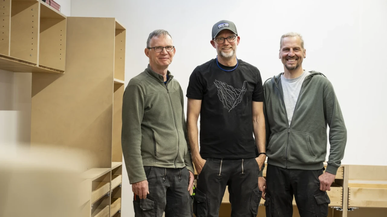 Drei stehende M&auml;nner l&auml;chelnd vor einem Regalsystem aus hellbraunem Holz. Der linke tr&auml;gt eine olivgr&uuml;ne Jacke, der mittlere ein schwarzes T-Shirt und eine Kappe, der rechte eine graue Kapuzenjacke. Im Hintergrund sind Regale und Schubladen zu sehen.