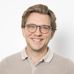 Mann mit blonden, leicht gewellten Haaren, hellblauer Brille und beigem Polohemd, l&auml;chelnd vor einem wei&szlig;en Hintergrund.