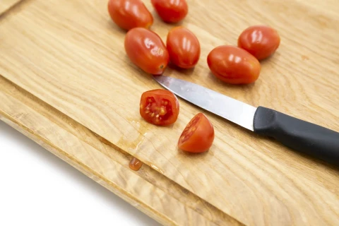 Kleine rote Cherrytomaten auf einem hellbraunen Holzschneidebrett, teilweise ganz, teilweise halbiert. Ein kleines Messer mit schwarzem Griff liegt neben den halbierten Tomaten.