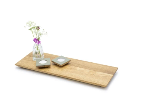 Holztablett aus hellem Holz mit einer schlichten Glasvase, die wei&szlig;e und violette Bl&uuml;ten enth&auml;lt. Daneben zwei quadratische Kerzenhalter aus grauem Material mit eingesetzten wei&szlig;en Teelichtern. Hintergrund in Wei&szlig;.
