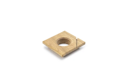 Quadratische Holzplatte aus hellbraunem Holz mit glatter Oberfl&auml;che. In der Mitte befindet sich ein rundes, vollst&auml;ndig durchgehendes Loch. Auf der rechten Seite ist ein schmaler, senkrechter Schlitz eingearbeitet.