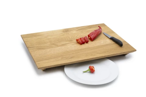 Holzschneidebrett mit erh&ouml;hten Beinen, darauf eine halbierte rote Chili, teilweise in Scheiben geschnitten. Schwarzes K&uuml;chenmesser rechts neben der Chili. Darunter ein wei&szlig;er Teller mit einer ganzen roten Chili.