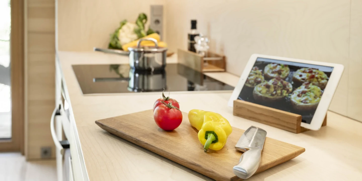 Holzschneidebrett mit einer roten Tomate, einer gelben Paprika und einem silbernen Messer, platziert auf einer hellen K&uuml;chenarbeitsplatte. Im Hintergrund ein Kochfeld mit einem Topf, ein Tablet mit ge&ouml;ffnetem Kochrezept auf einem Holzst&auml;nder und weitere K&uuml;chenutensilien.