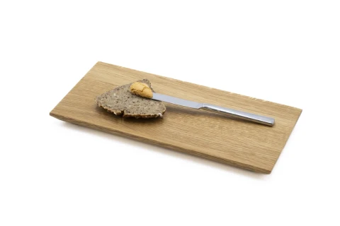 Scheibe dunkles K&ouml;rnerbrot mit einem Klecks Erdnussbutter darauf, neben einem silbernen Buttermesser auf einem rechteckigen Holzbrett.