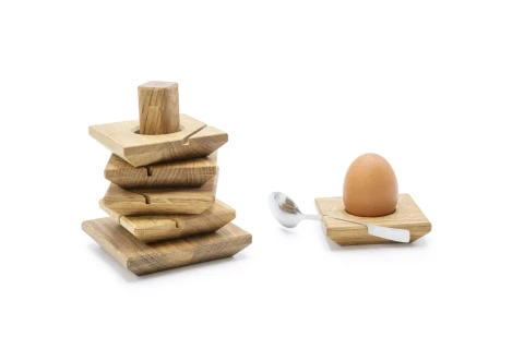 Holzstapel aus lose aufeinandergelegten, verschieden gro&szlig;en quadratischen Holzplatten auf der linken Seite, rechts ein schlichtes hellbraunes Ei in einem quadratischen Holz-Eierbecher, daneben ein silberner L&ouml;ffel. Erkennbar vor wei&szlig;em Hintergrund.