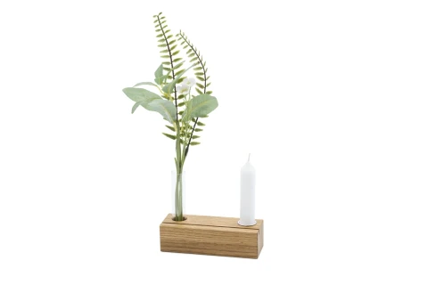 Holzblock mit zwei Aussparungen, einer f&uuml;r eine schmale Glasvase mit k&uuml;nstlichen gr&uuml;nen Zweigen und Bl&auml;ttern, die andere f&uuml;r eine wei&szlig;e, schlanke Kerze. Minimalistisches Design auf wei&szlig;em Hintergrund.