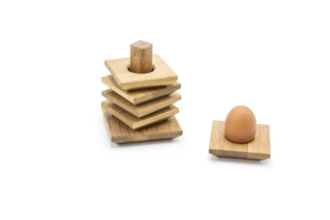 Holzspielzeug in Form eines Turms mit quadratischen Holzbl&ouml;cken auf einer Holzstange links. Rechts ein braunes Ei auf einer quadratischen Holzunterlage, beides vor wei&szlig;em Hintergrund.