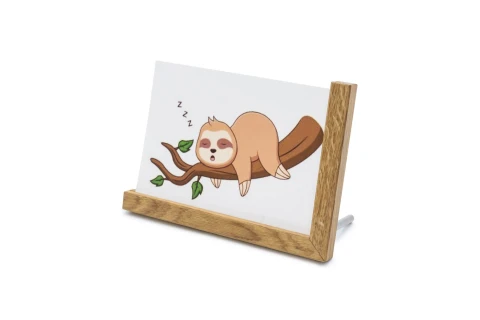 Schlafendes Faultier als Cartoon auf wei&szlig;em Hintergrund, liegend auf einem braunen Ast mit gr&uuml;nen Bl&auml;ttern, dar&uuml;ber drei Z-Zeichen als Schlafsymbol. Das Bild ist in einem Holzrahmen mit schr&auml;ger Standfl&auml;che eingefasst.