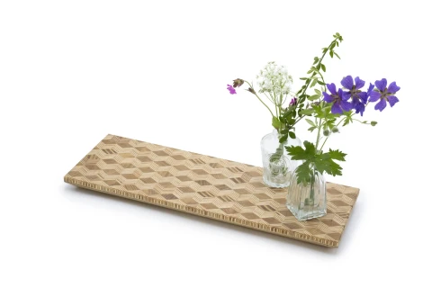 Holzbrett mit geometrischem W&uuml;rfelmuster aus hellem und dunklem Holz. Zwei kleine Glasvasen darauf, links mit wei&szlig;en Bl&uuml;ten und rechts mit lila Bl&uuml;ten und gr&uuml;nen Bl&auml;ttern.