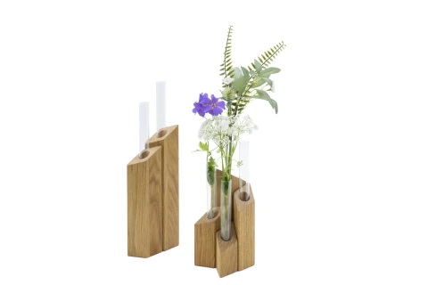 Zwei geometrische Holzvasen mit je drei eingelassenen, transparenten Glasr&ouml;hren. Die rechte Vase enth&auml;lt Blumen: eine violette Bl&uuml;te, wei&szlig;e Bl&uuml;ten und gr&uuml;ne Farn- und Bl&auml;tterzweige. Die linke Vase ist leer.
