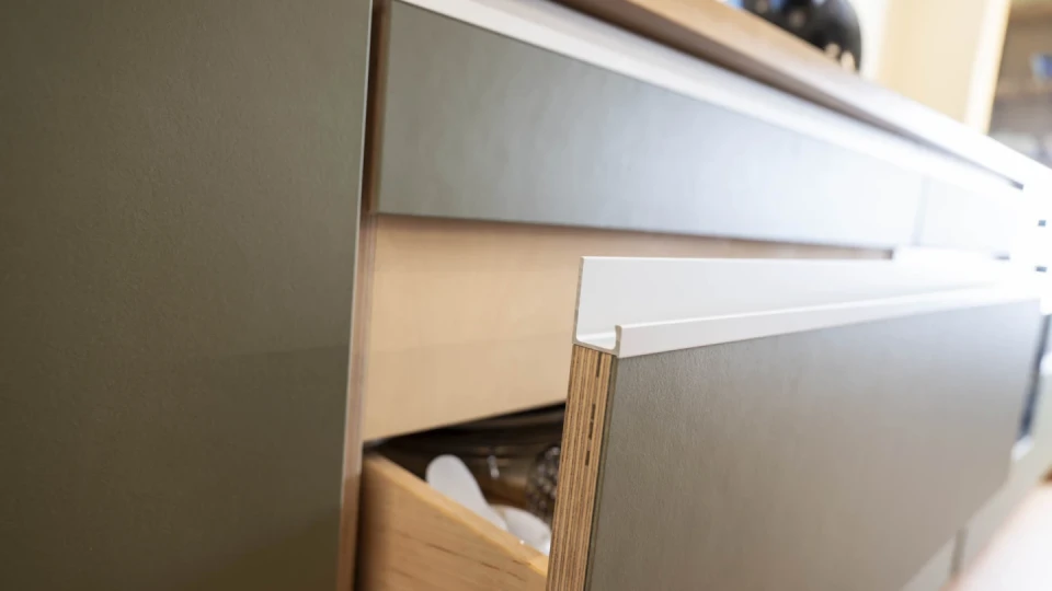 Gr&uuml;n-graue K&uuml;chenschr&auml;nke mit moderner Oberfl&auml;che, teilweise ge&ouml;ffnet, eine Holzschublade sichtbar, deren Inhalt Glas- und K&uuml;chenutensilien umfasst. Oben eine helle Holzarbeitsplatte mit einem schwarzen Wasserkocher im Hintergrund.