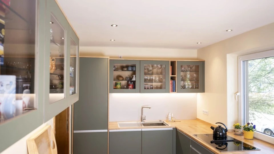 Moderne K&uuml;che mit gr&uuml;nen Einbauschr&auml;nken, teils mit Glast&uuml;ren, auf denen Geschirr sichtbar ist. Arbeitsplatten aus Holz, eine Sp&uuml;le mit Armatur, ein Kochfeld mit Wasserkocher und gelbem Blumentopf daneben. Rechts ein Fenster mit Blick auf gr&uuml;ne B&auml;ume. Deckenstrahler sorgen f&uuml;r Beleuchtung.